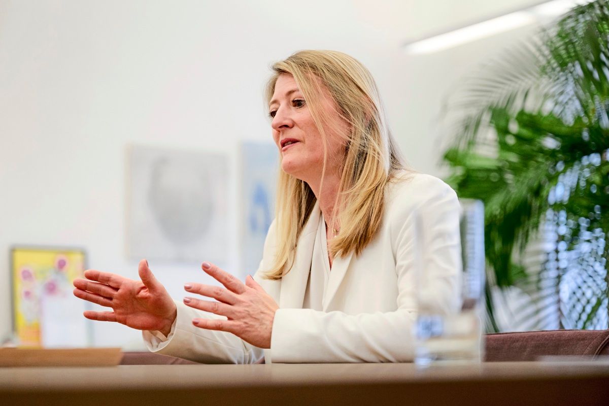 Eine Frau mit blondem Haar in einem weißen Blazer gestikuliert während eines Gesprächs. Im Hintergrund sind unscharfe Bilder und eine Pflanze erkennbar.