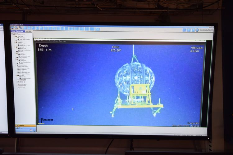 Ein Kamerabild auf einem Bildschirm zeigt einen kugelförmigen Detektor, der mit einem Kran hinabgelassen wird.