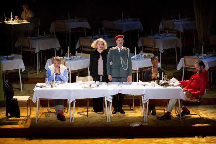 Theaterstück Reigen, Schauspielerinnen und Schauspieler sitzen bzw. stehen an einem langen Tisch.