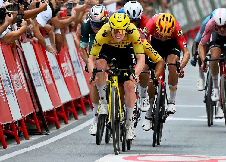 Ein Radrennfahrer in gelbem Trikot sprintet an der Spitze eines Feldes während der 11. Etappe der Vuelta a España 2026, umgeben von anderen Fahrern. Zuschauer stehen hinter Absperrungen und halten Kameras hoch.