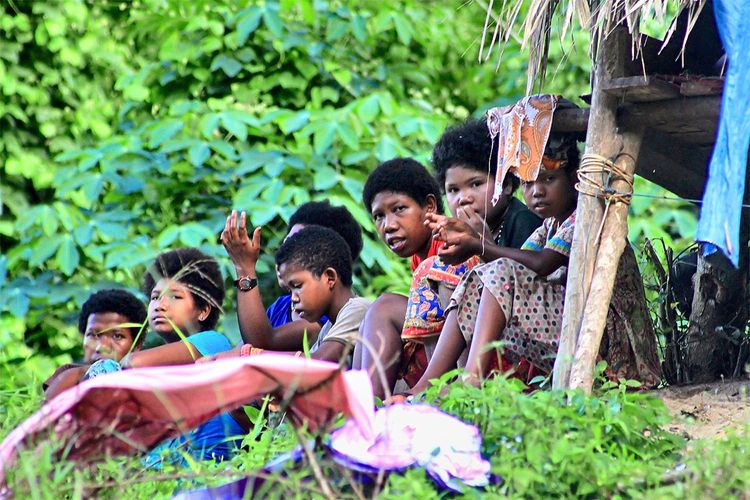 Eine Gruppe von Menschen, darunter auch Kinder, sitzt unter einem einfachen Unterstand in einer von üppigem Grün umgebenen Umgebung. Einige tragen bunte Kleidung, während sie miteinander interagieren. Im Vordergrund sind Pflanzen und Stoffe zu sehen.