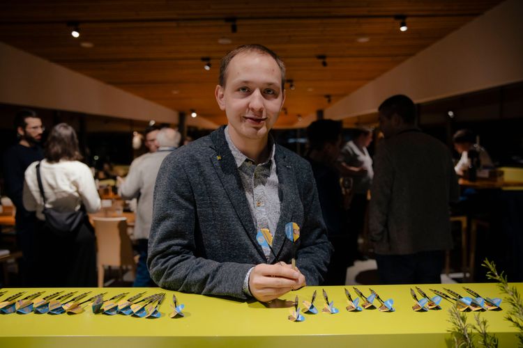 Florian Hofmann steht vor der Schank, auf der blau-gelb bedruckte Dartpfeile aufgereiht liegen