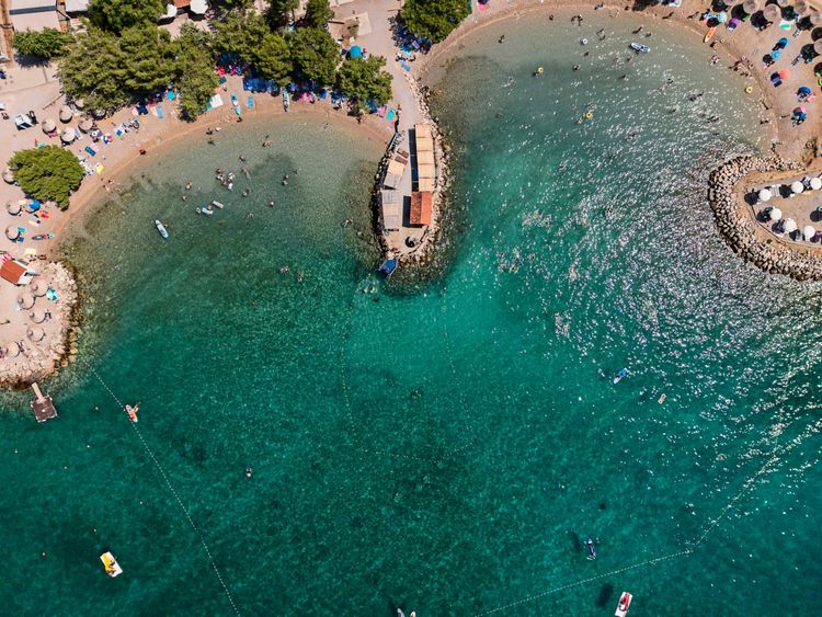 Drohnenaufnahme des Unterwasserparks Punat auf der Insel Krk, Kroatien. Der Strand ist von Bäumen umgeben, das Wasser ist klar, und Menschen genießen das Schwimmen und die Aktivitäten im Wasser. Sonnenschirme und Liegen säumen den Strand.