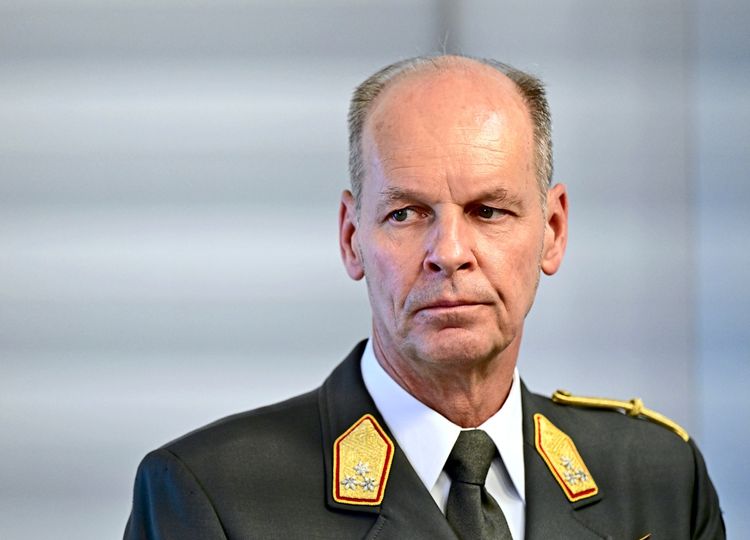 ALT-Text: Ein Mann in militärischer Uniform mit Abzeichen auf den Schultern. Er trägt ein weißes Hemd und eine schwarze Krawatte. Der Hintergrund ist neutral und unscharf.