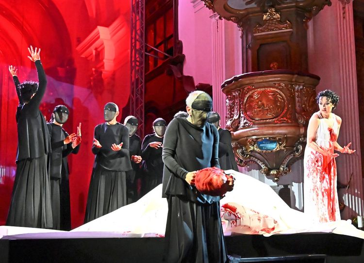 Eine Theaterinszenierung mit dramatischem Setting: Mehrere Personen in schwarzen Gewändern und Masken stehen auf der Bühne. Eine Person hält ein blutiges Objekt, während eine Frau in einem weißen, blutbefleckten Kleid agiert. Der Hintergrund zeigt eine prunkvolle, kirchenartige Architektur.