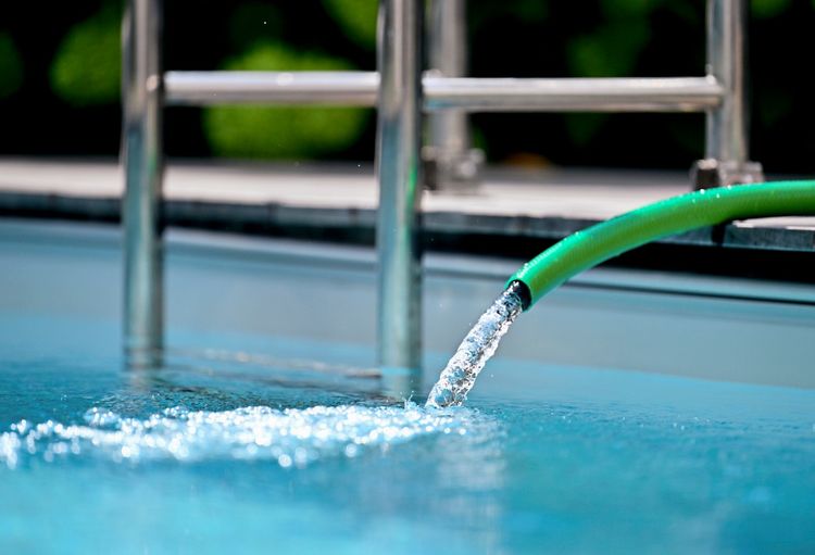 Ein grüner Wasserschlauch gibt Wasser in einen Swimmingpool ab. Im Hintergrund ist eine Metallleiter erkennbar.