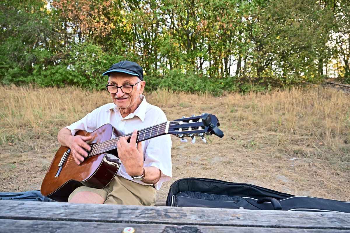 Ein älterer Mann mit Brille und Baseballkappe sitzt im Freien und spielt auf einer Akustikgitarre. Im Hintergrund ist eine trockene Wiese und Wald zu sehen. Neben ihm liegt eine geöffnete Gitarrentasche.