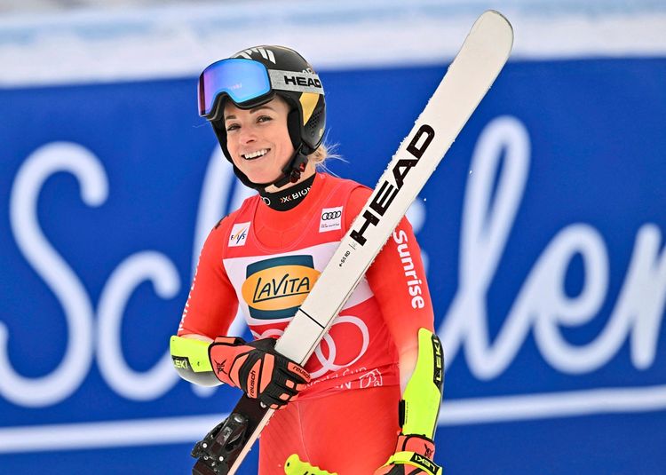 Eine Skirennläuferin in roter Rennkleidung hält einen Ski und lächelt. Sie trägt einen Helm mit Skibrille und Handschuhe. Im Hintergrund ist ein verschwommener blauer Schriftzug sichtbar.