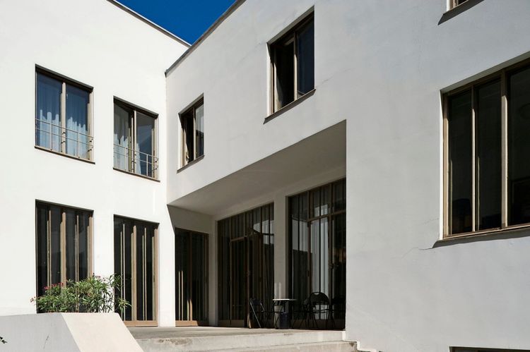 Ansicht eines modernen Gebäudes mit weißer Fassade und großen rechteckigen Fenstern, gestaltet im minimalistischen Stil. Das Gebäude verfügt über einen überdachten Eingangsbereich mit gläsernen Türen und schlichten metallenen Gartenmöbeln. Klarer, geometrischer Stil.