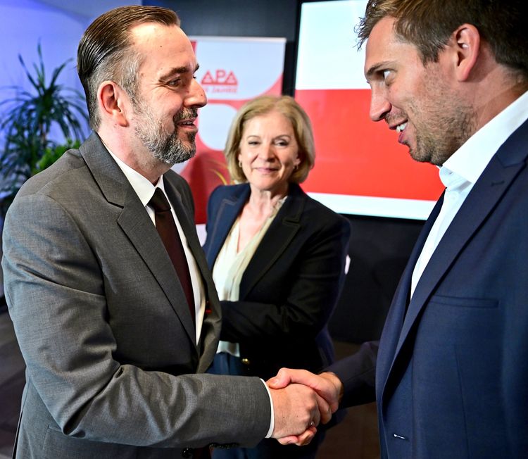 Drei Personen bei einem formellen Anlass: Links ein Mann im Anzug, der einem anderen Mann rechts die Hand schüttelt. In der Mitte steht eine Frau im Anzug und lächelt. Hintergrund mit rotem Schriftzug und Pflanzen. Veranstaltung anlässlich 