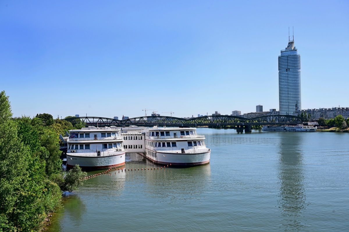 Zwei große Schiffe liegen auf dem Wasser eines breiten Flusses vor Anker. Im Hintergrund ist eine Brücke sowie ein hoher, moderner Turm erkennbar. Bäume und weitere Gebäude säumen die Umgebung unter klarem, blauem Himmel.