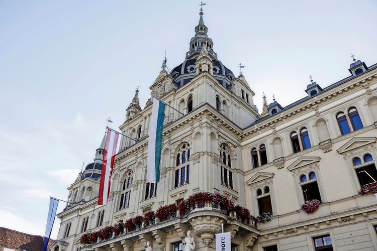 Ansicht des Grazer Rathauses mit historischen Fassadendetails, Fahnen und Blumenschmuck an den Balkonen, aufgenommen an einem sonnigen Tag mit klarem Himmel.