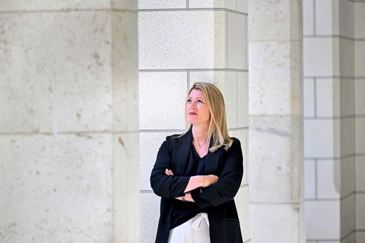 Eine Frau mit blonden Haaren steht zwischen massiven Steinsäulen und schaut nachdenklich nach oben. Sie trägt einen schwarzen Blazer und hat die Arme verschränkt.
