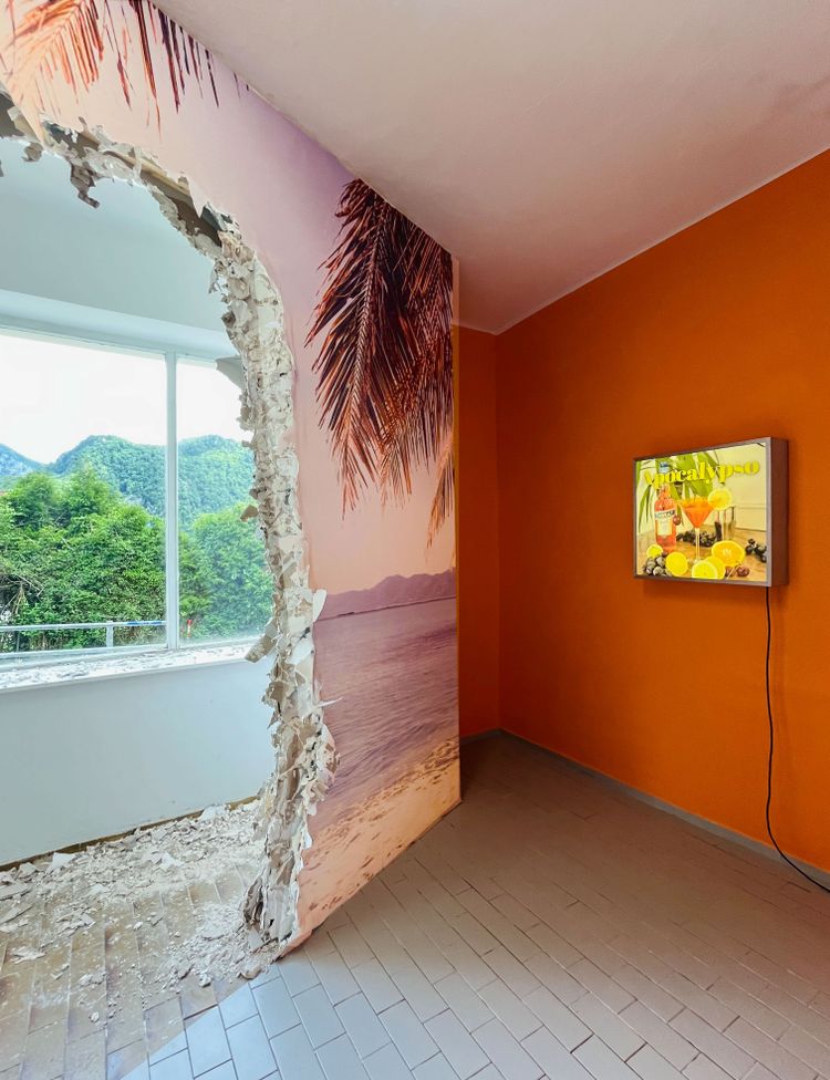 Rechts eine orange gestrichene Wand mit Bildschirm, auf dem tropische Früchte und ein Cocktail zu sehen sind, Aufschrift 