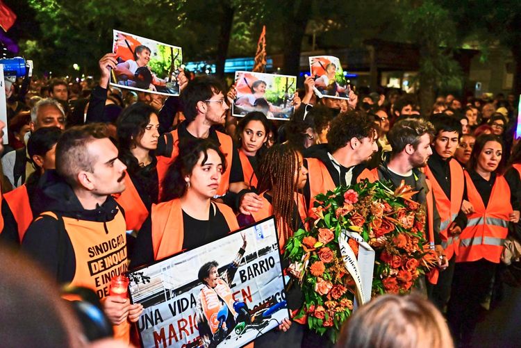 Eine Gruppe von Menschen, teils in orangefarbenen Warnwesten, nimmt an einem Gedenkmarsch in Madrid teil. Einige halten Plakate mit Fotos, andere tragen einen Kranz aus orangefarbenen Blumen. Die Veranstaltung erinnert an Maricarmen, eine Frau, die nach einer Zwangsräumung gestorben ist und als Symbol für den Kampf für Wohnrechte gilt.