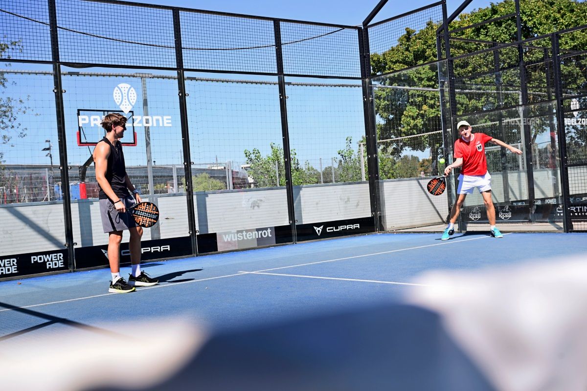 Zwei Männer spielen auf einem Padel-Tennisplatz, umgeben von einem Gitterzaun. Einer trägt ein schwarzes Outfit, der andere ein rotes Shirt und eine weiße Kappe. Im Hintergrund sind Bäume und ein Gebäude zu sehen.
