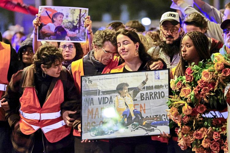 Eine Gruppe von Demonstrierenden marschiert in Madrid, Spanien, mit emotionalen Gesichtsausdrücken. Sie tragen orangefarbene Warnwesten, halten Fotos und Porträts einer älteren Frau sowie Blumenschmuck. Die Demonstration erinnert an eine verstorbene Frau, deren Zwangsräumung Wohnungsproteste ausgelöst hat.