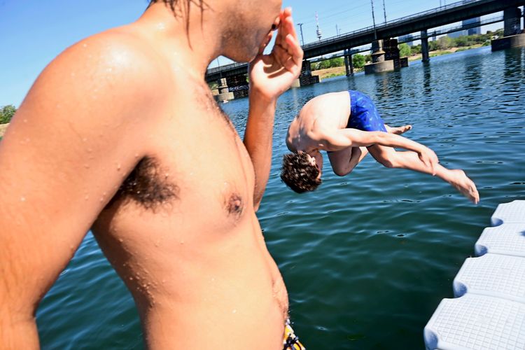 Ein Mann steht im Vordergrund, während ein anderer Mann im Hintergrund von einem schwimmenden Steg ins Wasser springt. Im Hintergrund sind eine Brücke und Bäume zu sehen. Es ist ein sonniger Sommertag.