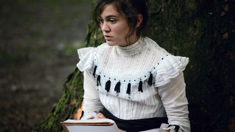 Milena Straube als Luise von Kummerveldt sitzt in einem weißen Kleid vor einem Baum und hält ein Buch in der Hand.