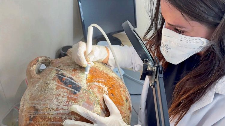 Eine Person in Schutzkleidung und Mundschutz reinigt sorgfältig eine alte Amphore mit einem kleinen Werkzeug unter einer beleuchteten Lupe.