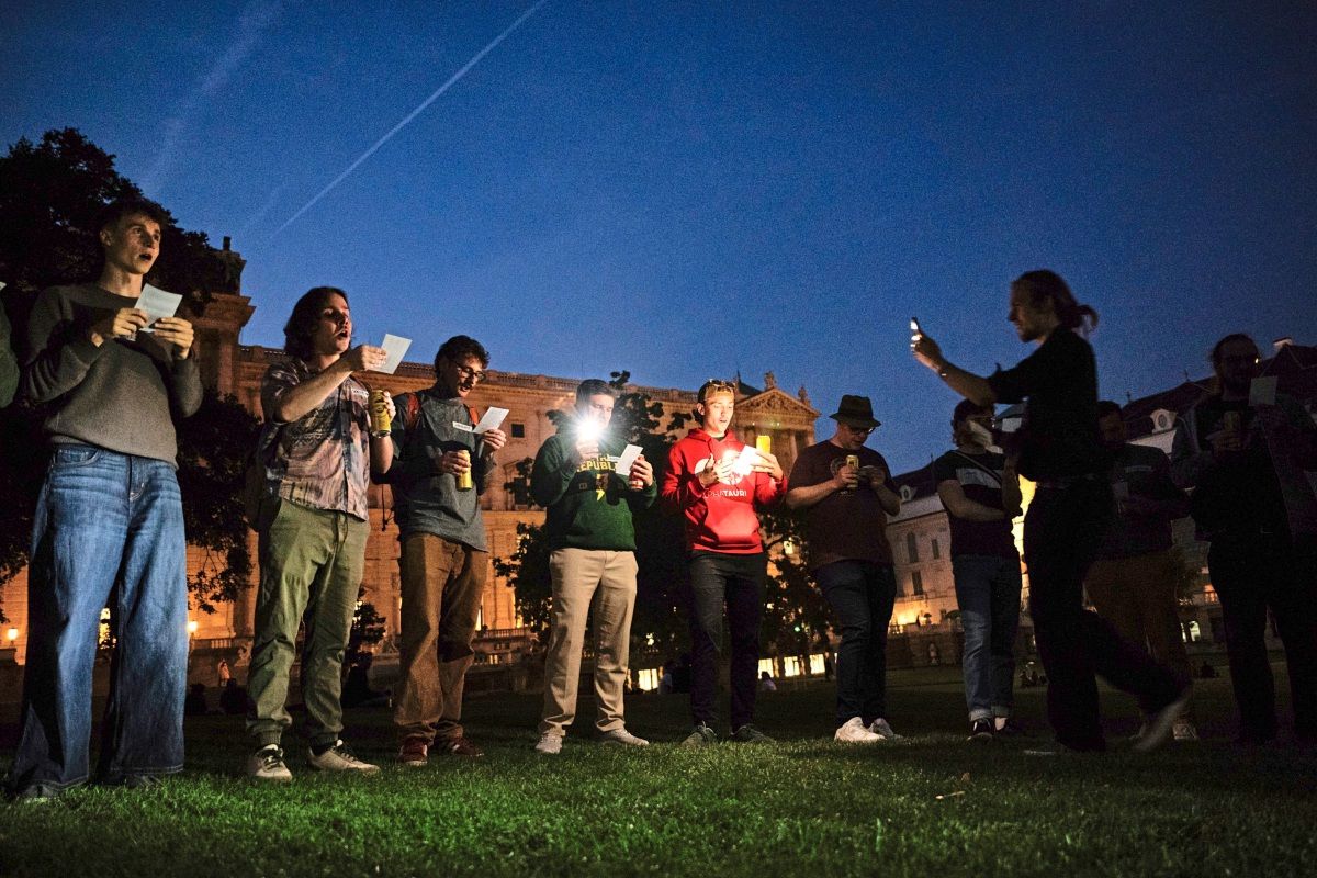 Eine Gruppe von Menschen steht nachts im Freien auf einer Wiese. Einige halten Zettel in den Händen, während andere ihre Smartphones als Lichtquelle nutzen. Im Hintergrund sind beleuchtete historische Gebäude zu sehen.