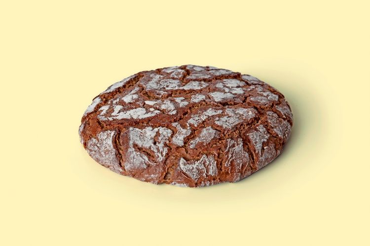 Ein rundes Roggenbrot mit rustikaler Kruste und Mehl bestäubt, auf einem hellgelben Hintergrund.