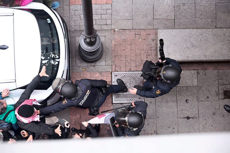 Blick von oben auf eine Auseinandersetzung zwischen Polizisten und Demonstranten während einer Kundgebung vor der Regionalregierung von Madrid. Die Polizei blockiert den Weg von Protestierenden, die versuchen, ein Gebäude zu betreten. Einige der Demonstranten tragen farbige Kleidung und Schals. Im Hintergrund ist ein geparktes Auto zu sehen.