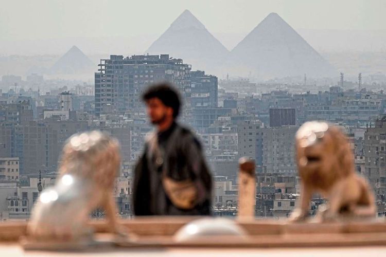 Im Hintergrund sieht man die Cheops-, Chephren- und Menkheres-Pyramiden, dann moderne Bauten der Stadt Gizeh, im Vordergrund unscharf einen Mann sowie zwei Löwenstatuen.