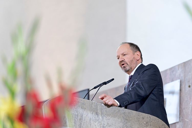 Ein Mann im Anzug spricht an einem Rednerpult in einer Kirche. Im Vordergrund sind unscharfe Blumen zu sehen.