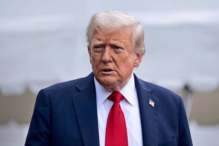 Ein älterer Mann mit weißem Haar trägt einen dunkelblauen Anzug, ein weißes Hemd und eine rote Krawatte. An seinem Revers ist ein Ansteckpin mit einer US-Flagge zu sehen. Der Hintergrund ist unscharf und zeigt weiße Strukturen.