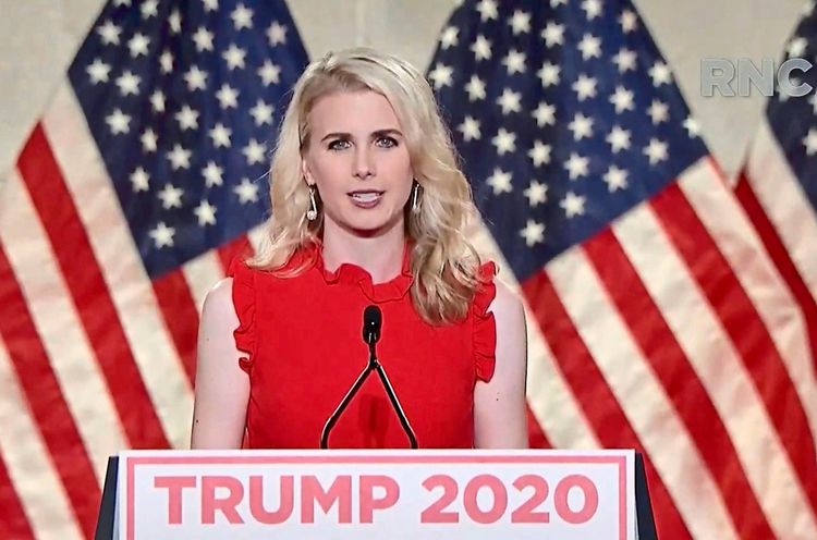 Eine Frau mit blondem Haar steht vor mehreren US-amerikanischen Flaggen und spricht in ein Mikrofon. Sie trägt ein rotes Kleid und steht hinter einem Pult mit der Aufschrift 