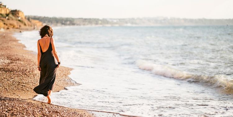 Eine Frau in einem langen schwarzen Kleid spaziert barfuß am sandigen und steinigen Strand entlang, während Wellen das Ufer erreichen. Im Hintergrund ist die Küstenlinie sichtbar.