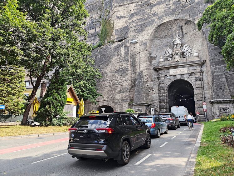 Eine Straßenszene in Salzburg, die den Eingang des historischen Sigmundstors (Neutor) im Mönchsberg zeigt. Autos stehen in einer Schlange vor dem Tunnel, darunter ein Taxi. Links sind Bäume und ein Gebäude mit gelben Sonnenschirmen zu sehen. Rechts führt ein Radfahrer am Straßenrand entlang.