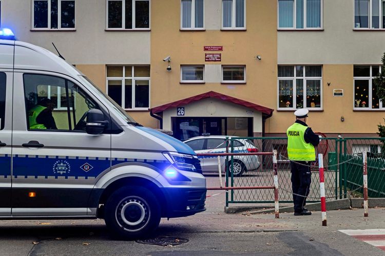 Ein Polizeiwagen mit aktivem Blaulicht steht vor einem Schulgebäude mit der Hausnummer 26. Ein Polizist in Uniform und gelber Warnweste steht neben einer Absperrung in der Nähe des Eingangs der Schule. Im Hintergrund ist das Schulgebäude mit Fenstern und einem Vordach zu sehen.