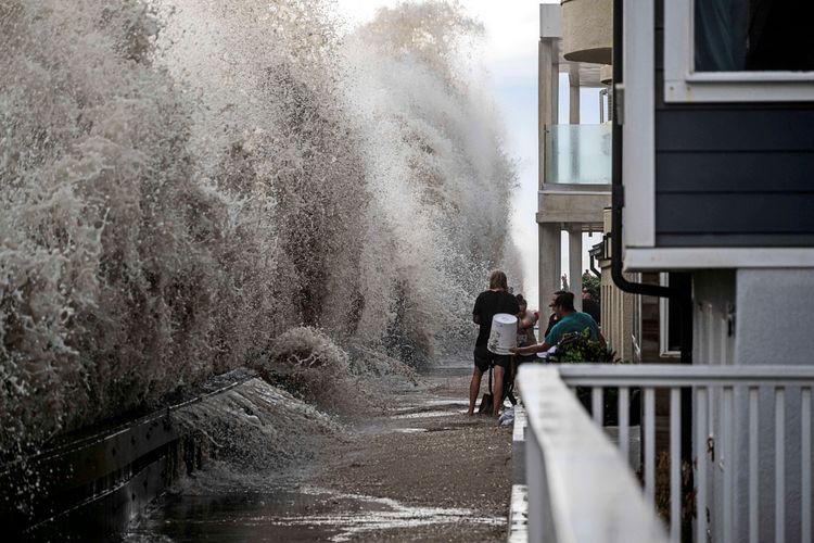 Links spritzt graues Meerwasser senkrecht vor einer Promenade mehrere Meter in die Höhe, rechts sieht man Häuserfassaden, vor denen Menschen stehen und mit Eimern Wasser hinausschöpfen.