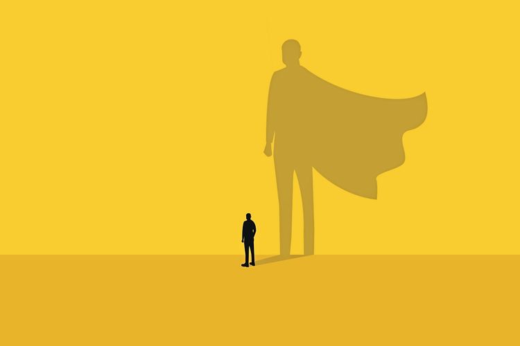 Ein kleiner Mann steht auf einem gelben Hintergrund, während sein Schatten die Silhouette eines Superhelden mit wehenden Umhangs darstellt. Symbol für Erfolg, Führung und Teamarbeit.