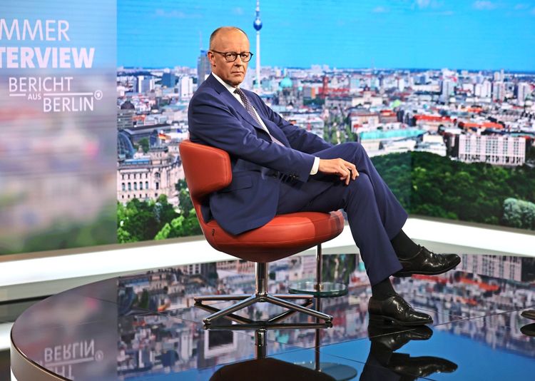 Ein Mann in einem blauen Anzug sitzt auf einem roten Stuhl in einem Fernsehstudio. Im Hintergrund ist eine Panoramasicht von Berlin mit dem Fernsehturm zu sehen. Text auf der Studiowand lautet „Sommerinterview – Bericht aus Berlin“.