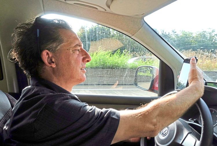 Ein Mann mittleren Alters sitzt am Steuer eines Autos und schaut während der Fahrt auf sein Mobiltelefon. Der Innenraum des Fahrzeugs ist sichtbar, ebenso wie ein Blick nach draußen auf eine grüne Landschaft. Die Situation deutet auf Ablenkung und ein potenzielles Unfallrisiko hin.