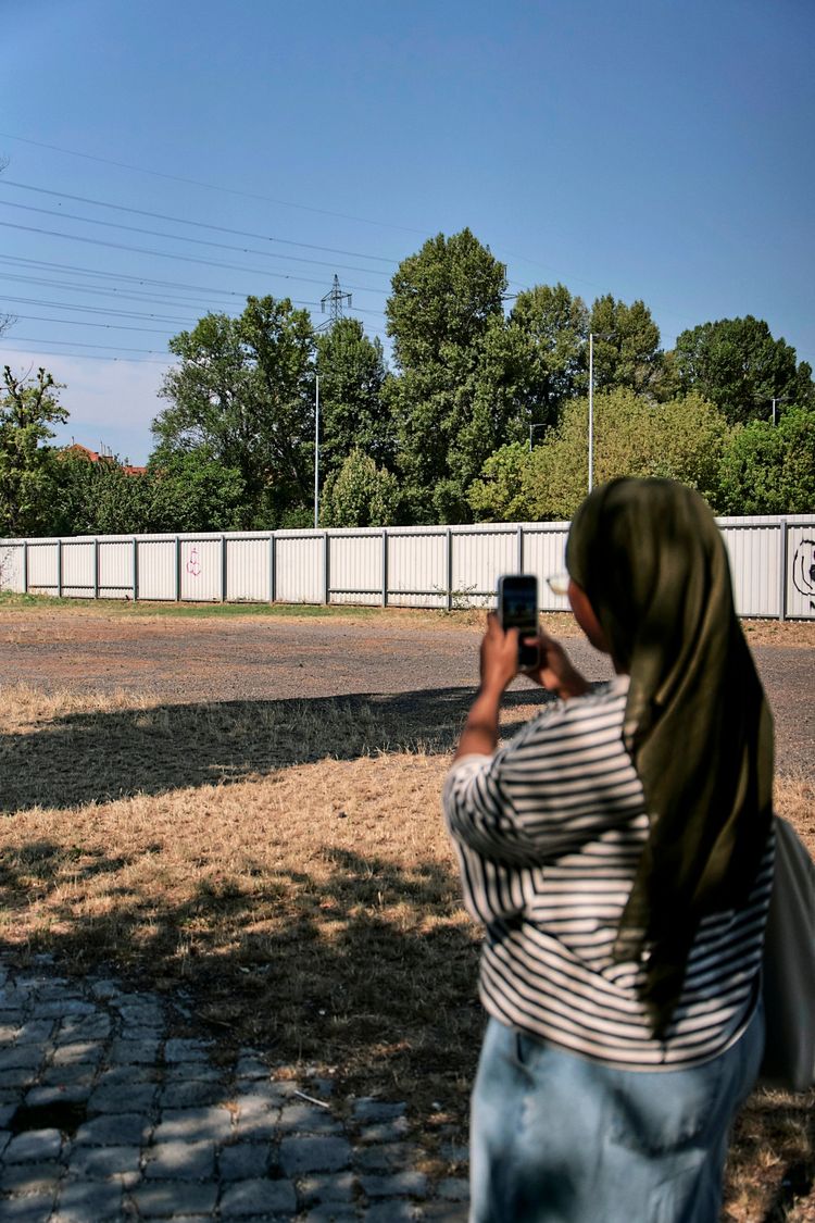 Eine Person mit gestreiftem Oberteil und grünem Kopftuch steht auf einer Wiese und fotografiert mit ihrem Smartphone. Im Hintergrund sind eine Mauer, Bäume und Stromleitungen zu sehen. Die Szene spielt an einem sonnigen Tag.