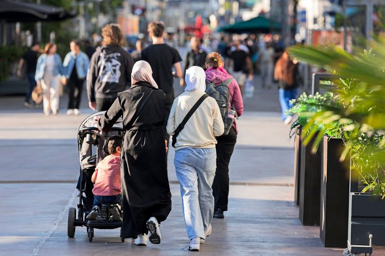 In der Fußgängerzone Favoritenstraße in Wien spazieren Menschen. Im Vordergrund schieben zwei Frauen mit Kopftüchern einen Kinderwagen. Im Hintergrund sind weitere Personen und Geschäfte zu sehen, die die belebte Atmosphäre widerspiegeln.