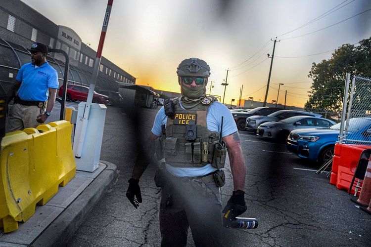 Ein ICE-Beamter (Immigration and Customs Enforcement) mit Schutzweste und Helm hält eine Pfefferspray-Dose vor dem Delaney Hall Gefängniszentrum in Newark, New Jersey. Im Hintergrund ist eine Absperrung, geparkte Autos und ein Sonnenuntergang zu sehen.