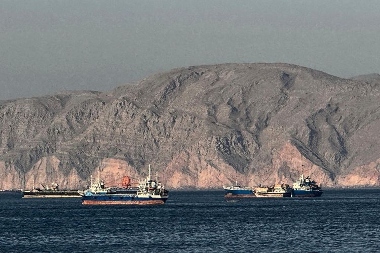 Mehrere Schiffe sind im Wasser der Straße von Hormus vor Anker gelegt, mit der felsigen Küstenlandschaft der Halbinsel Musandam im Norden Omans im Hintergrund.