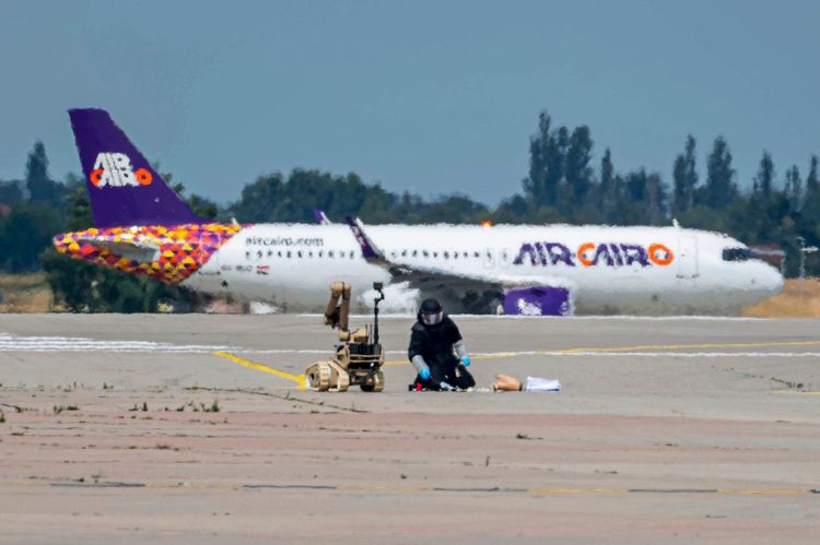 Ein Flughafen mit einem Flugzeug der Fluggesellschaft Air Cairo im Hintergrund. Im Vordergrund arbeitet eine Person in Schutzkleidung auf dem Boden nahe einem Roboter, der offenbar zur Untersuchung eines Objekts verwendet wird. Die Szene deutet auf einen Sicherheitsvorfall hin.