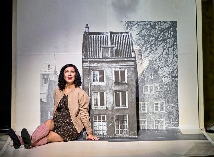 Eine Frau sitzt auf dem Boden vor einer schwarz-weißen Fotografie eines mehrstöckigen Gebäudes. Sie trägt ein gepunktetes Kleid, eine beige Strickjacke, rosa Kniestrümpfe und schwarze Schuhe, während sie nach oben schaut.