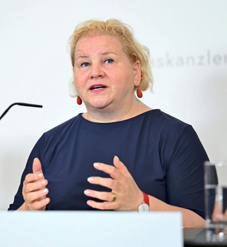 Eine Frau mit blonden Haaren und blauer Kleidung spricht gestikulierend während einer Pressekonferenz im Bundeskanzleramt in Wien. Im Hintergrund ist eine weiße Wand mit verschwommenem Schriftzug erkennbar.