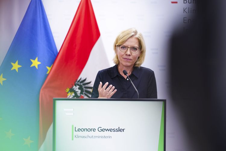 Ministerin Leonore Gewessler bei einer Pressekonferenz, sprechend hinter einem Pult. Hinter ihr eine Österreich-Flagge sowie eine der EU.