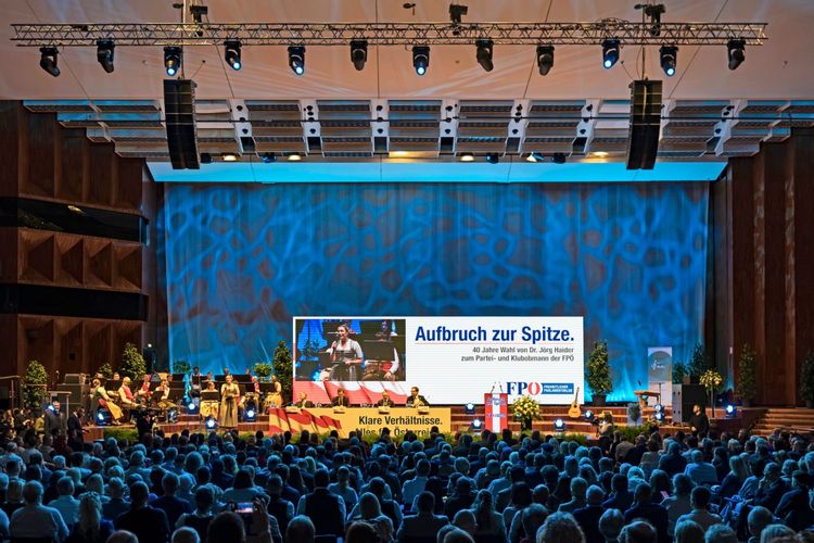 Eine Veranstaltung in einem großen Saal zum 40. Jahrestag der Wahl von Jörg Haider zum Parteiobmann der FPÖ in Innsbruck. Auf der Bühne befinden sich Redner, Musiker und große Bildschirme mit Schriftzügen wie „Aufbruch zur Spitze“. Im Saal sitzt ein großes Publikum, die Bühne ist beleuchtet.