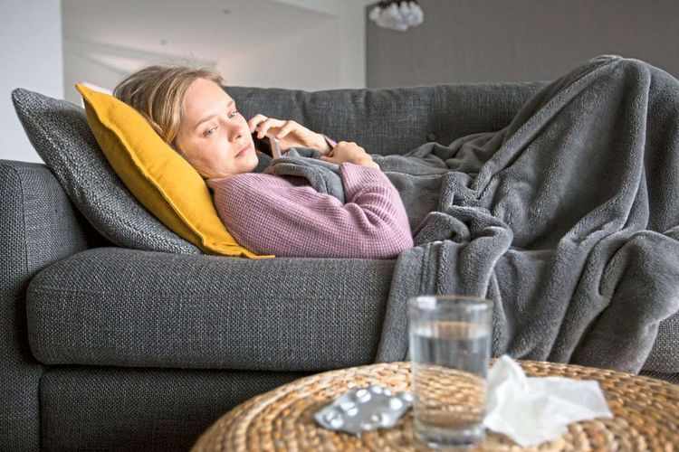 Eine offensichtlich erkrankte Frau, mit Medikamenten auf dem Tisch vor sich, liegt auf einer Couch, das Handy am Ohr