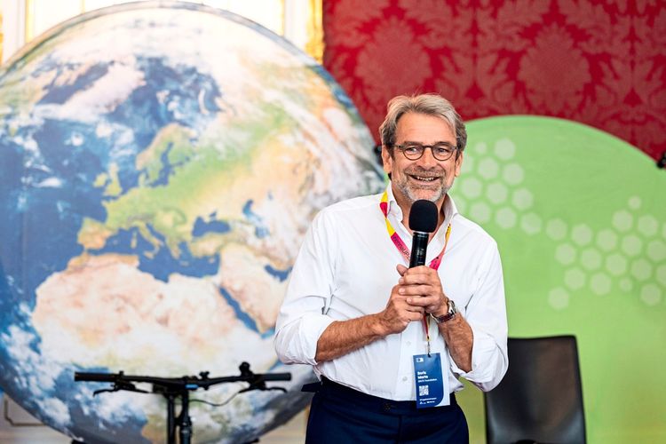 Ein Mann mit Brille und Mikrofon in der Hand steht lächelnd vor einem großen Globus und einer farbigen Wand. Er trägt ein weißes Hemd und ein Namensschild.