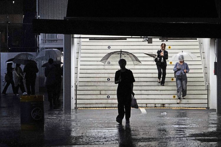 Personen verlassen bei starkem Regen eine Bahnhofstreppe in Tokio. Einige tragen Regenschirme, während Wasser auf dem Boden sichtbar ist. Die Szene wirkt düster mit dunkler Beleuchtung.